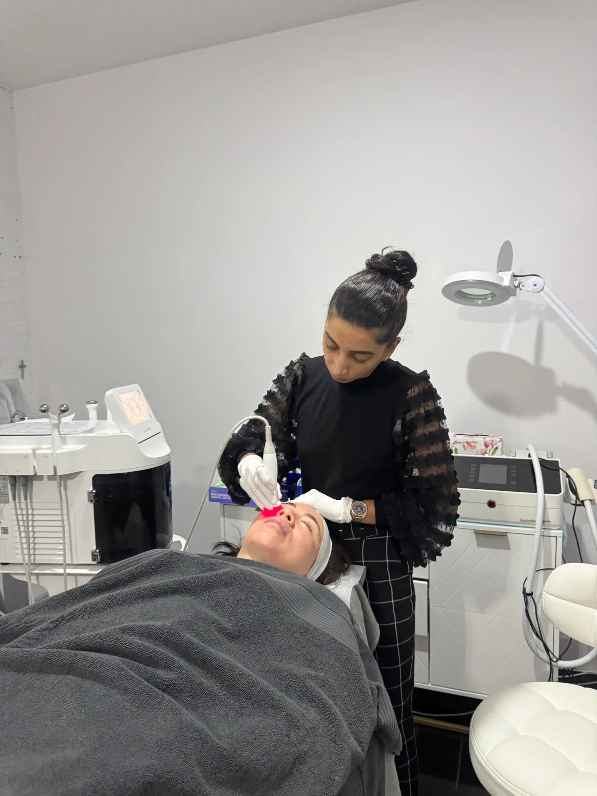 In a well-equipped treatment room, Anushka is carefully administering a skin resurfacing procedure to a client who is comfortably draped with a grey blanket.