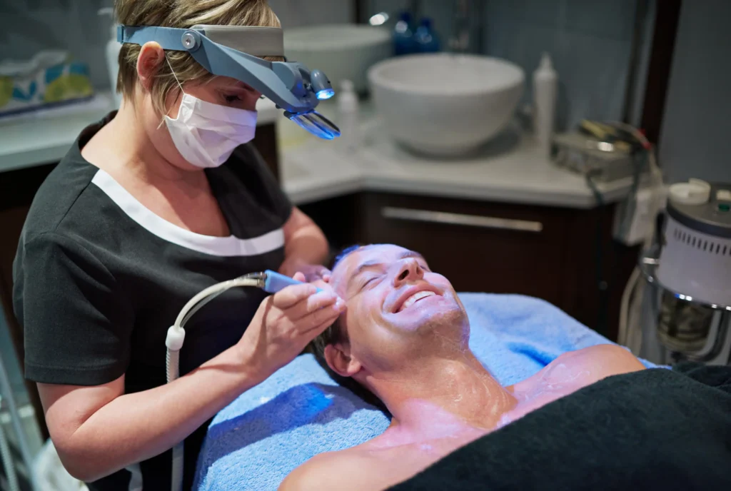 The image shows a smiling client receiving an Aqua Pure Facial, a 3-in-1 treatment for cleansing, hydrating, and rejuvenating the skin.